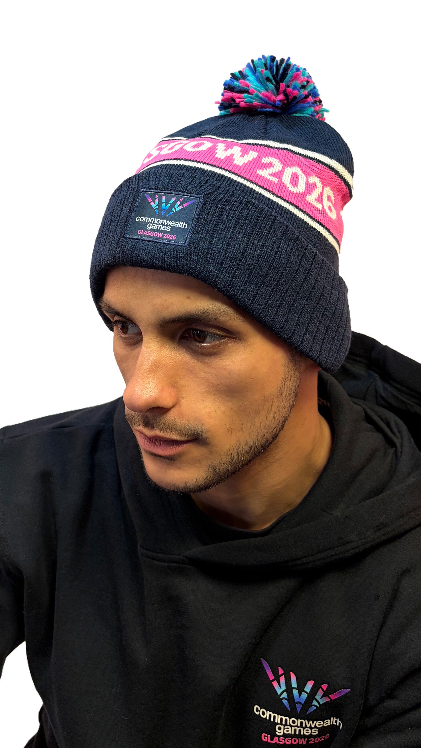 Front view of man wearing navy beanie with multi-colour pom pom and Glasgow 2026 on a pink stripe and Glasgow 2026 logo.