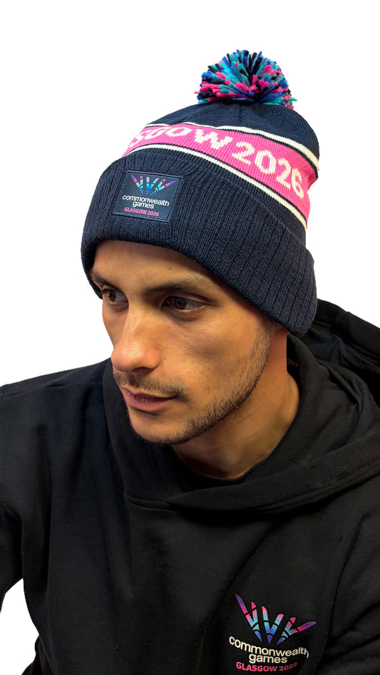 Front view of man wearing navy beanie with multi-colour pom pom and Glasgow 2026 on a pink stripe and Glasgow 2026 logo.