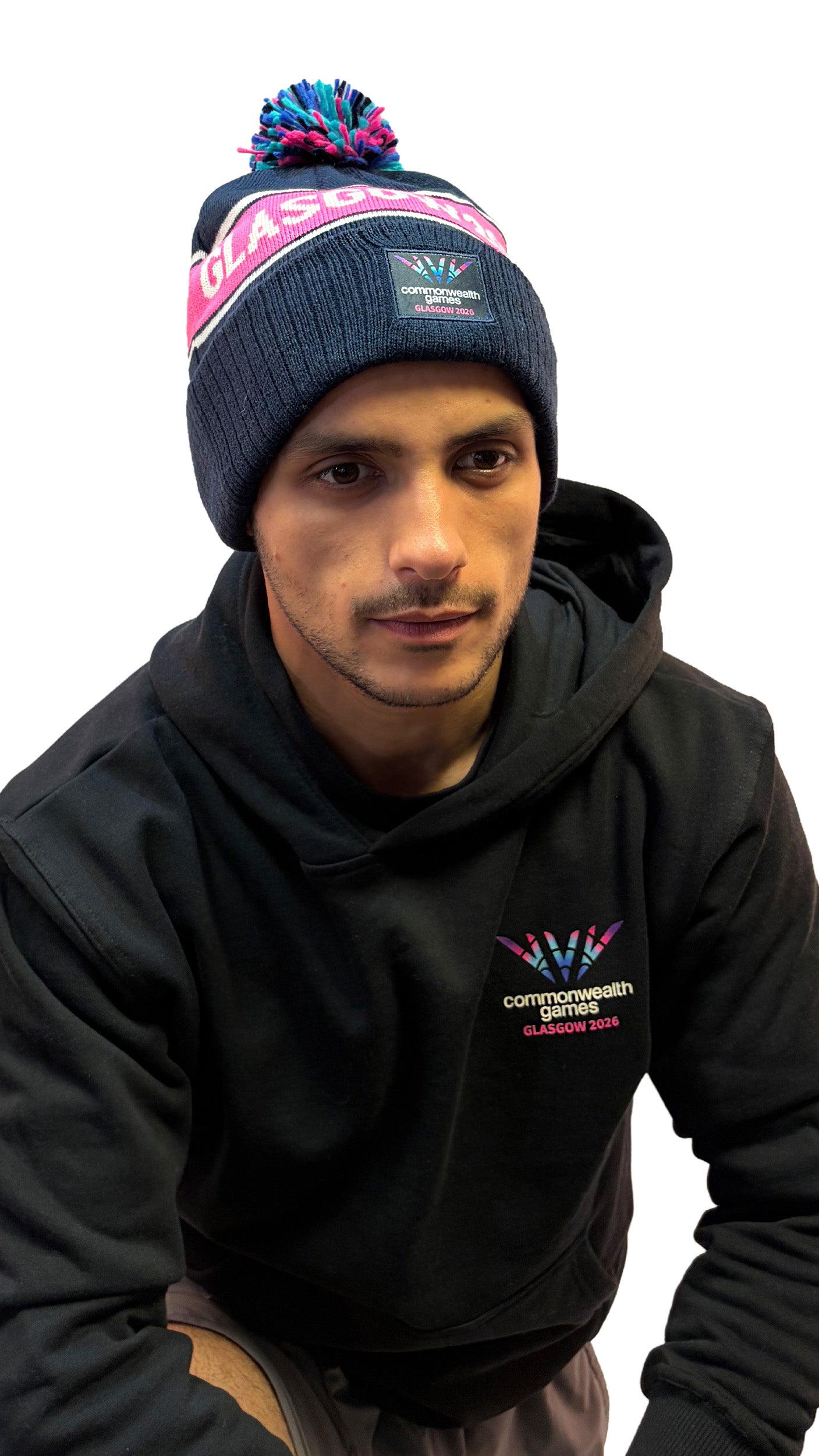 Front view of man wearing navy beanie with multi-colour pom pom and Glasgow 2026 on a pink stripe and Glasgow 2026 logo.