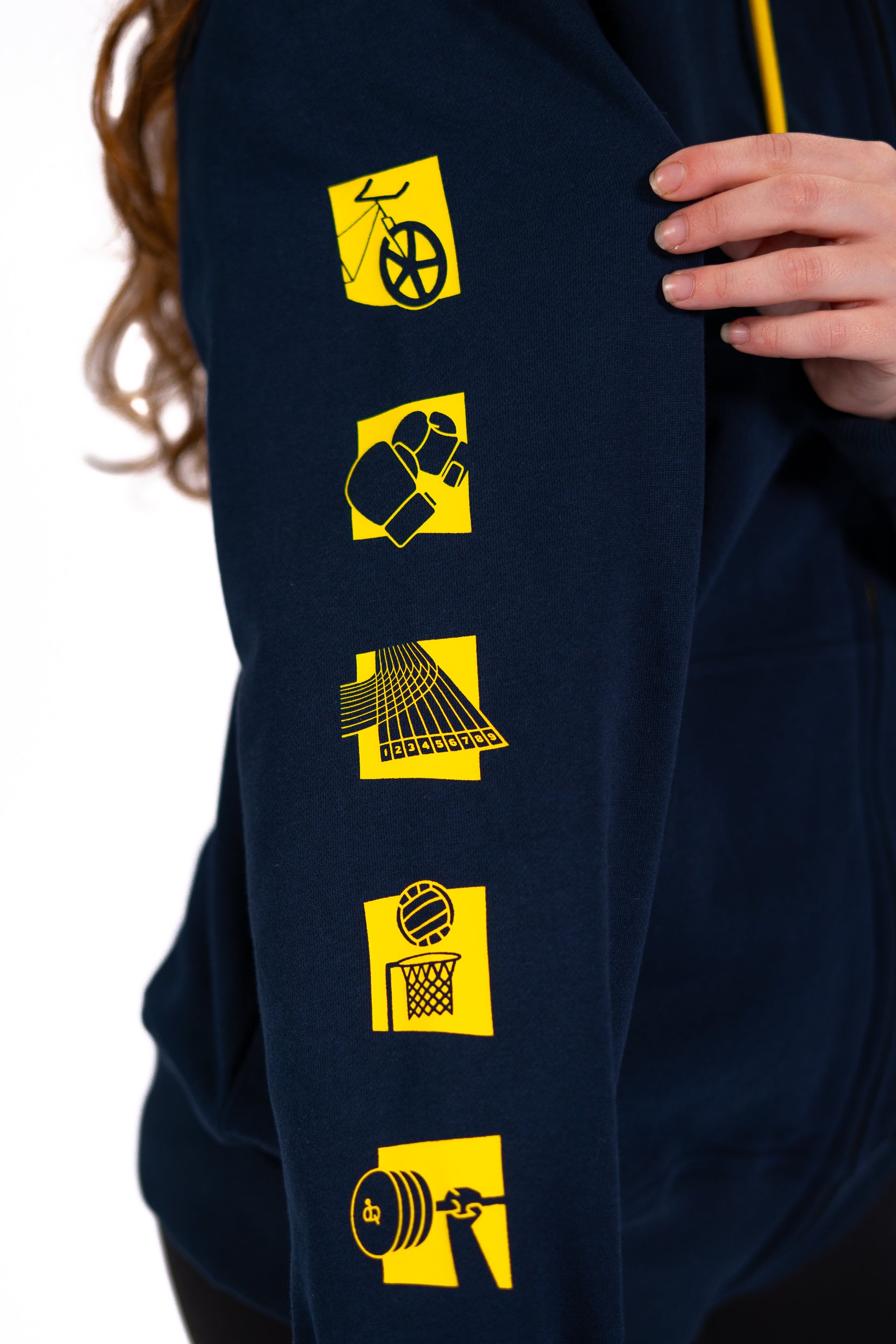 Close up of woman wearing contrast navy/yellow Glasgow 2026 Pictograms hoodie showing sport pictograms on sleeve.