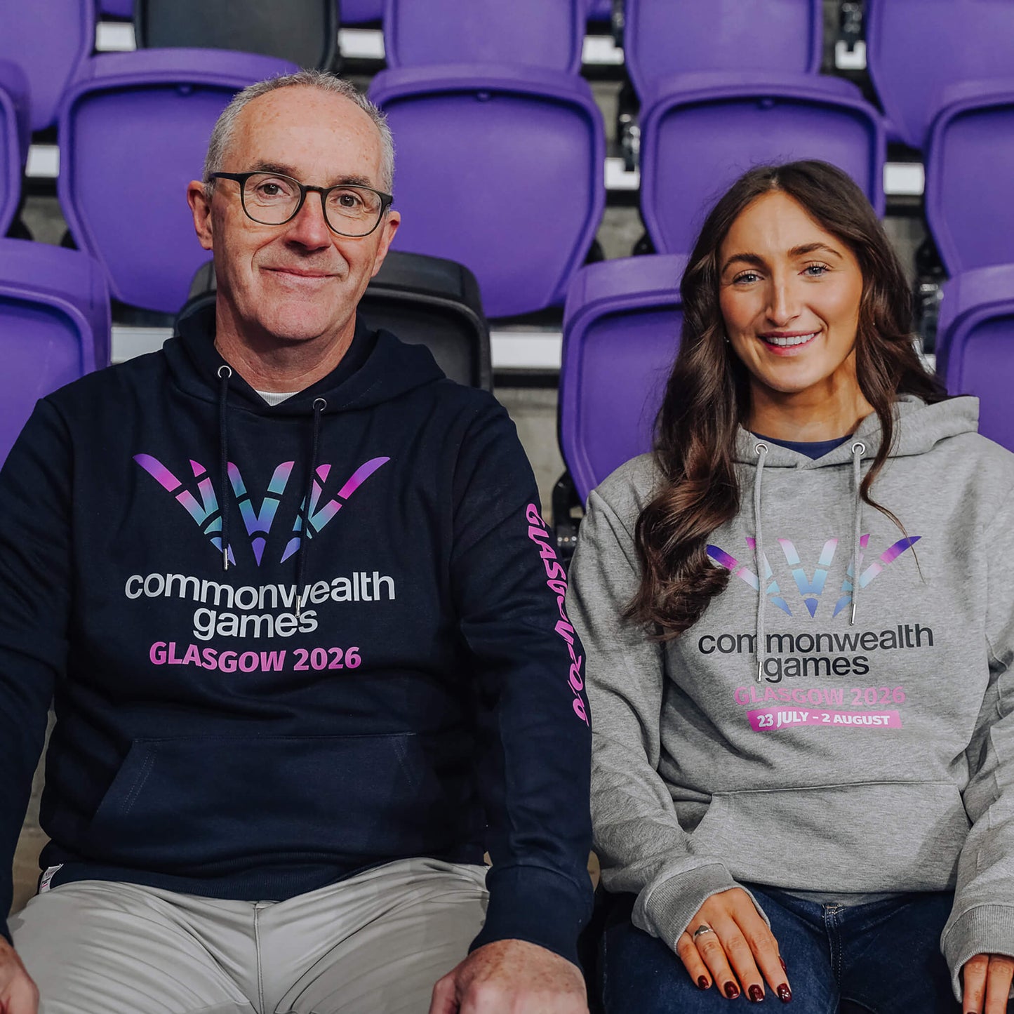Man wearing navy Glasgow 2026 hoodie accompanied by woman wearing grey Glasgow 2026 hoodie.