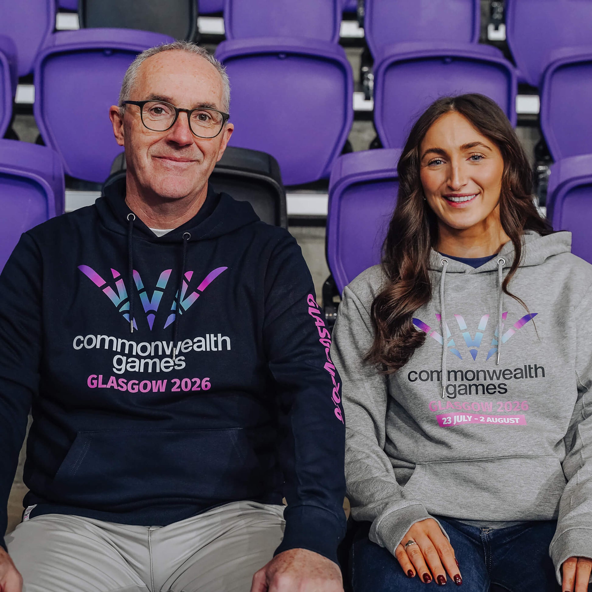 Man wearing navy Glasgow 2026 hoodie accompanied by woman wearing grey Glasgow 2026 hoodie.